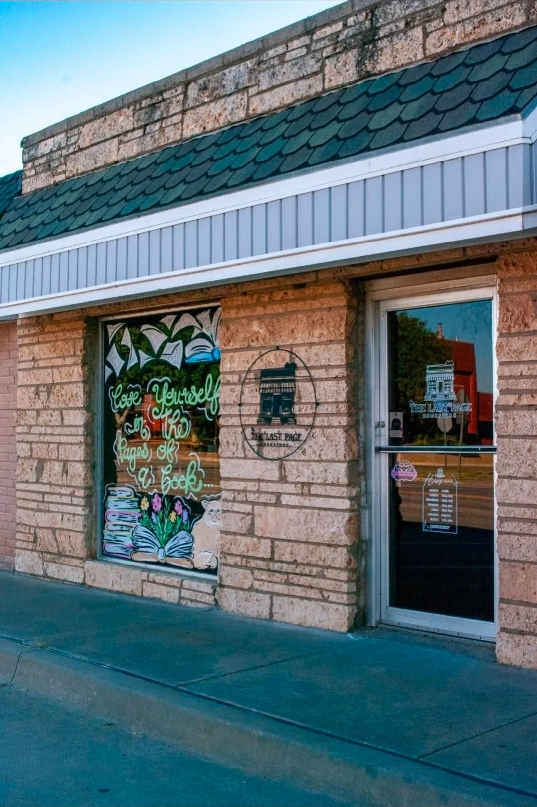 The storefront has a large window with colorful artwork that says "Love Yourself in the Pages of a Book" and depicts an open book with flowers, a cat, and a stack of books. To the right, there is a glass door and the sign "The Last Page Bookstore" on the window and door.