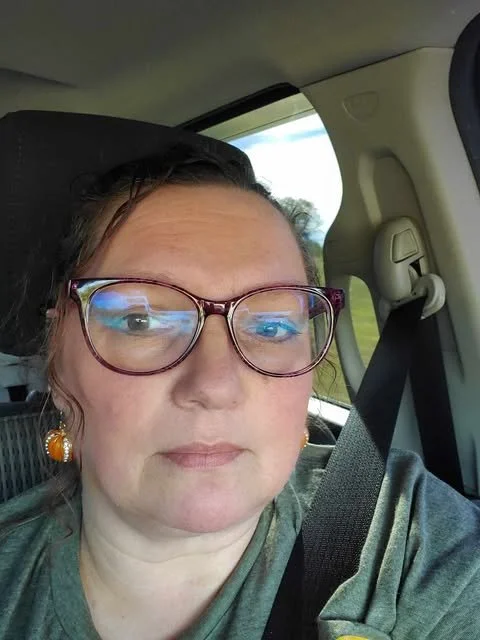 A woman with glasses and earrings sitting in a car, wearing a grey shirt, with a seatbelt on, and a window showing a green landscape outside.