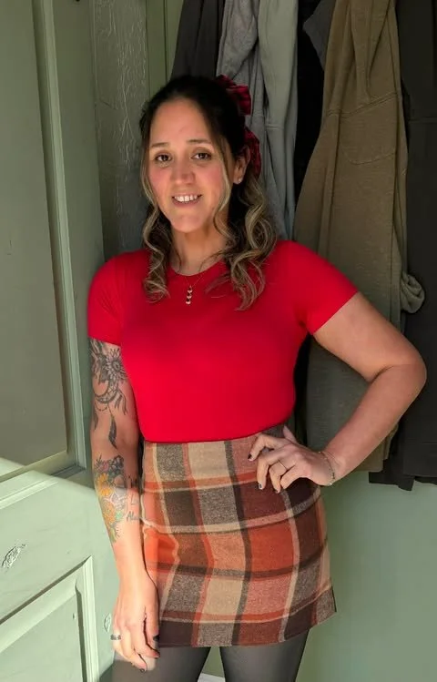 A woman with wavy blonde hair wearing a red top, a plaid skirt, and tights, standing in front of a closet with hanging clothes, smiling at the camera.