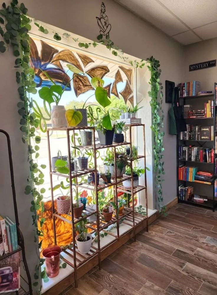 A window sill with numerous potted plants on a metal shelf, with a decorative cut-out design on the window, surrounded by green hanging plants, in a room with wooden flooring and a bookshelf filled with books.