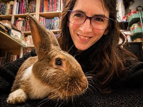 A woman with glasses smiling next to a brown and white rabbit in a room with bookshelves.