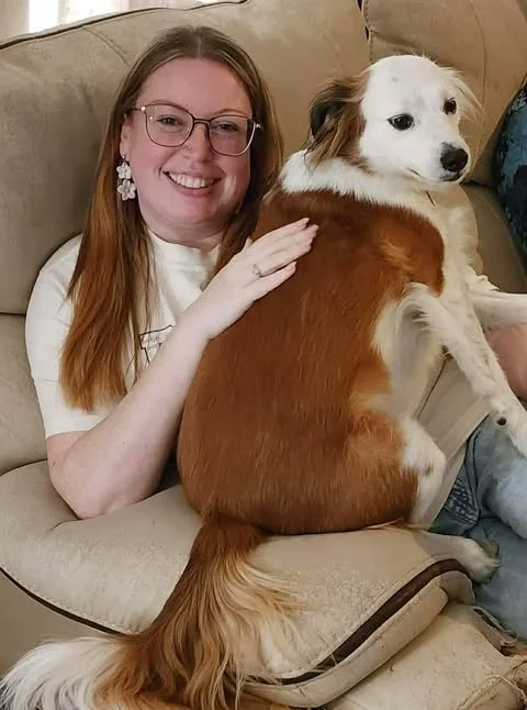 A smiling woman with glasses sitting on a beige couch, holding a large, long-haired dog on her lap.