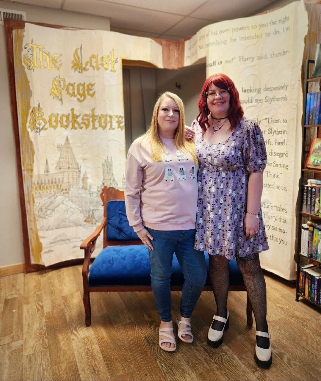 Two women standing next to each other in front of a Harry Potter-themed backdrop that reads "The Last Page of Hogwarts." They are inside a bookstore or library, with bookshelves on the right. One woman has blonde hair and is wearing a pink sweatshirt with cartoon characters, while the other has red hair and is wearing a purple floral dress and platforms.