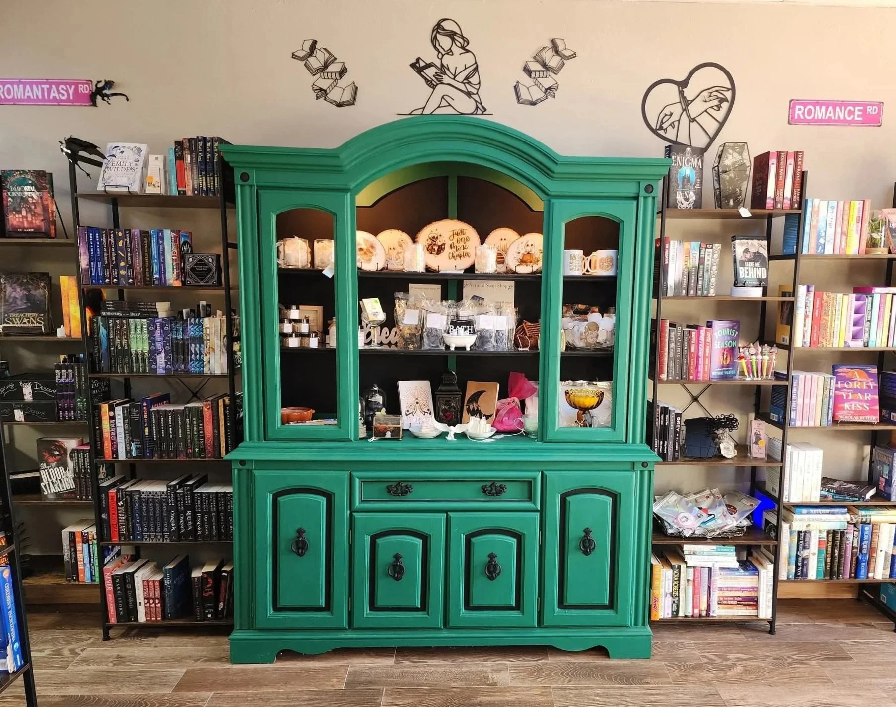 A bright green vintage-style china cabinet with curved details and ornate black hardware, displaying decorative ceramics and trinkets, set against a background of black metal bookcases filled with colorful books and wall art of books and reading-themed illustrations, including a girl reading and hearts with hands, in a bookstore or library.