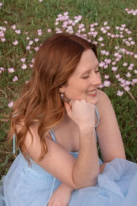 A woman with red hair, smiling with her eyes closed, sitting outdoors among pink flowers and green grass, resting her chin on her hand.