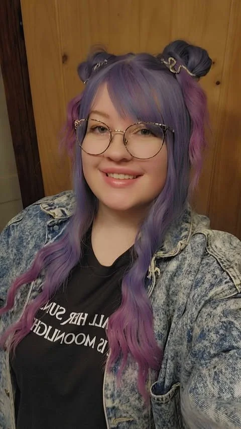 Young woman with colorful purple and blue hair styled in buns and loose waves, wearing glasses, a black T-shirt, and a denim jacket, smiling in front of a wooden wall.
