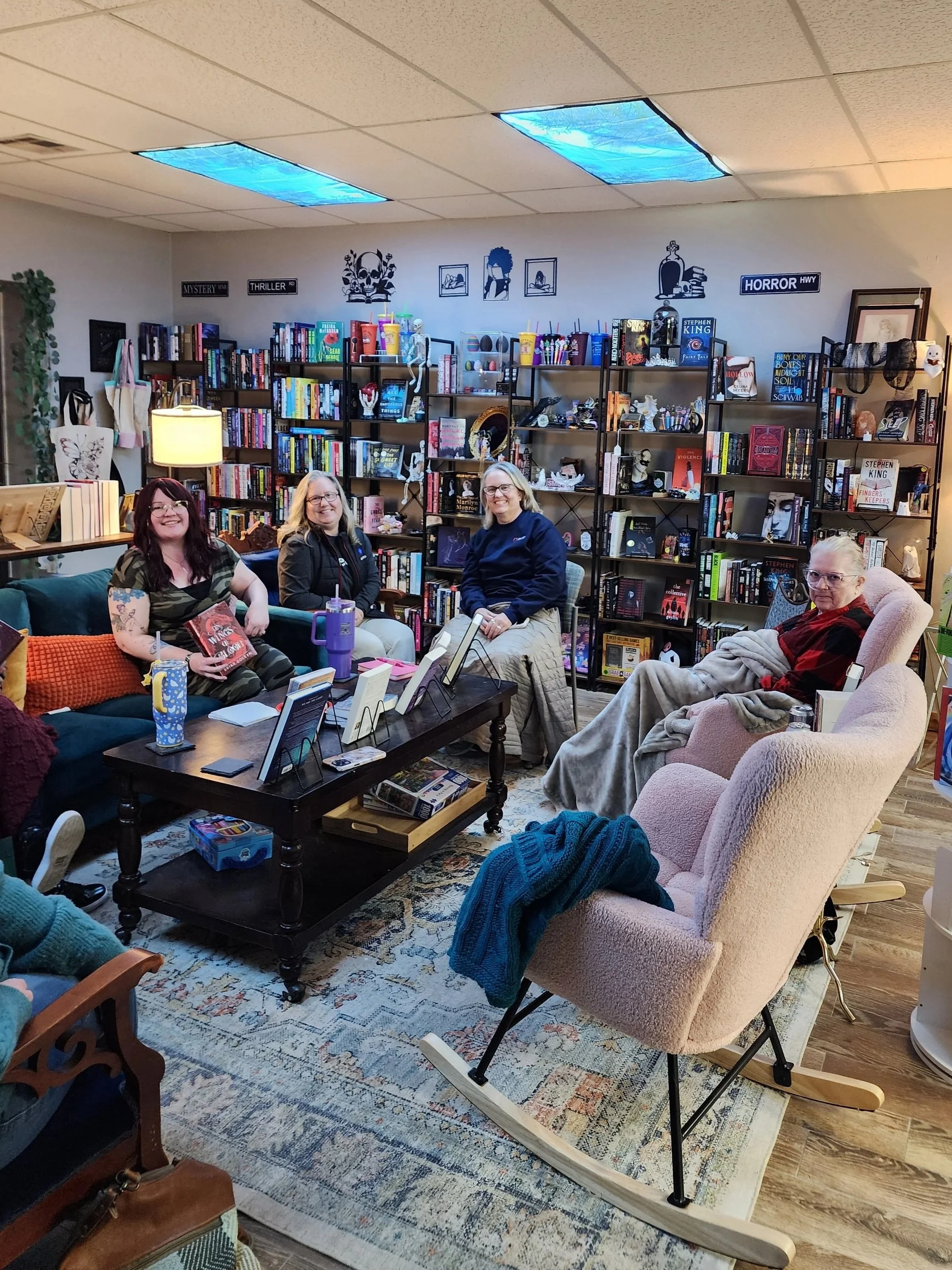 Four women sitting in a cozy room filled with bookshelves, books, and decorations, participating in a book club meeting or similar gathering.