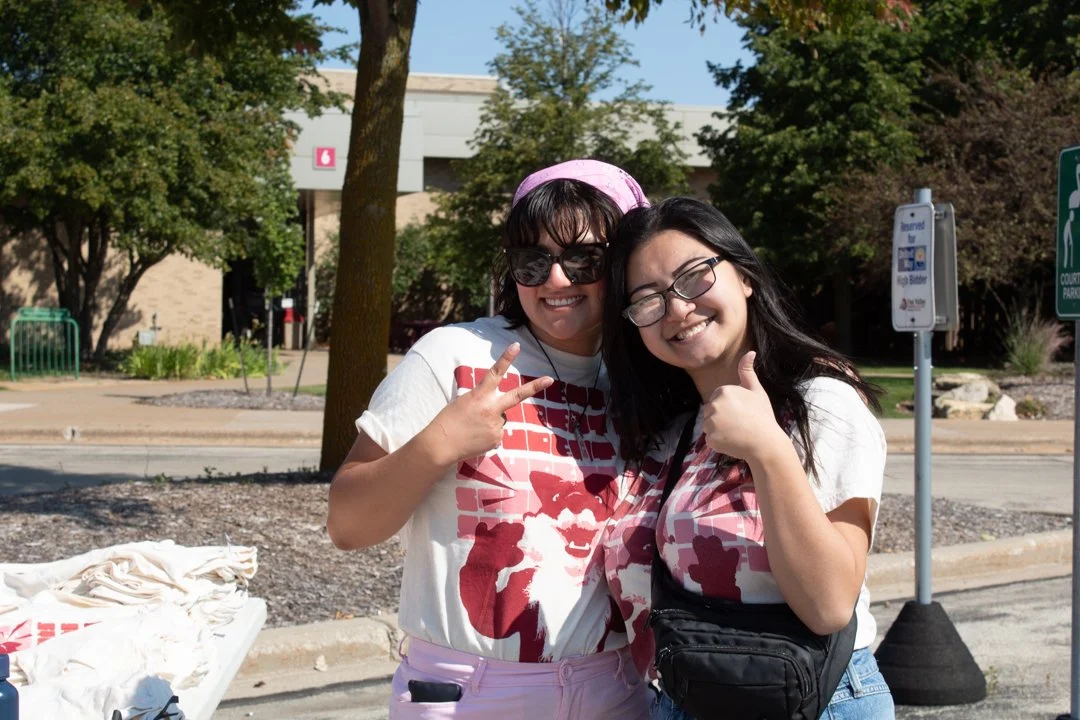 two female students wearing the student life t-shirt smiling at the camera, one with a thumbs up and another with a peace sign