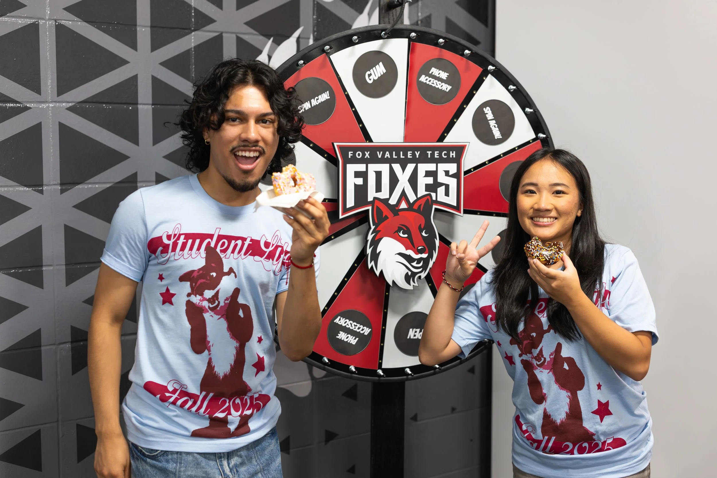 a male and female student smiling at the camera with a blue 2025 student life t-shirt. Both are holding donuts with a bite taken out of them.
