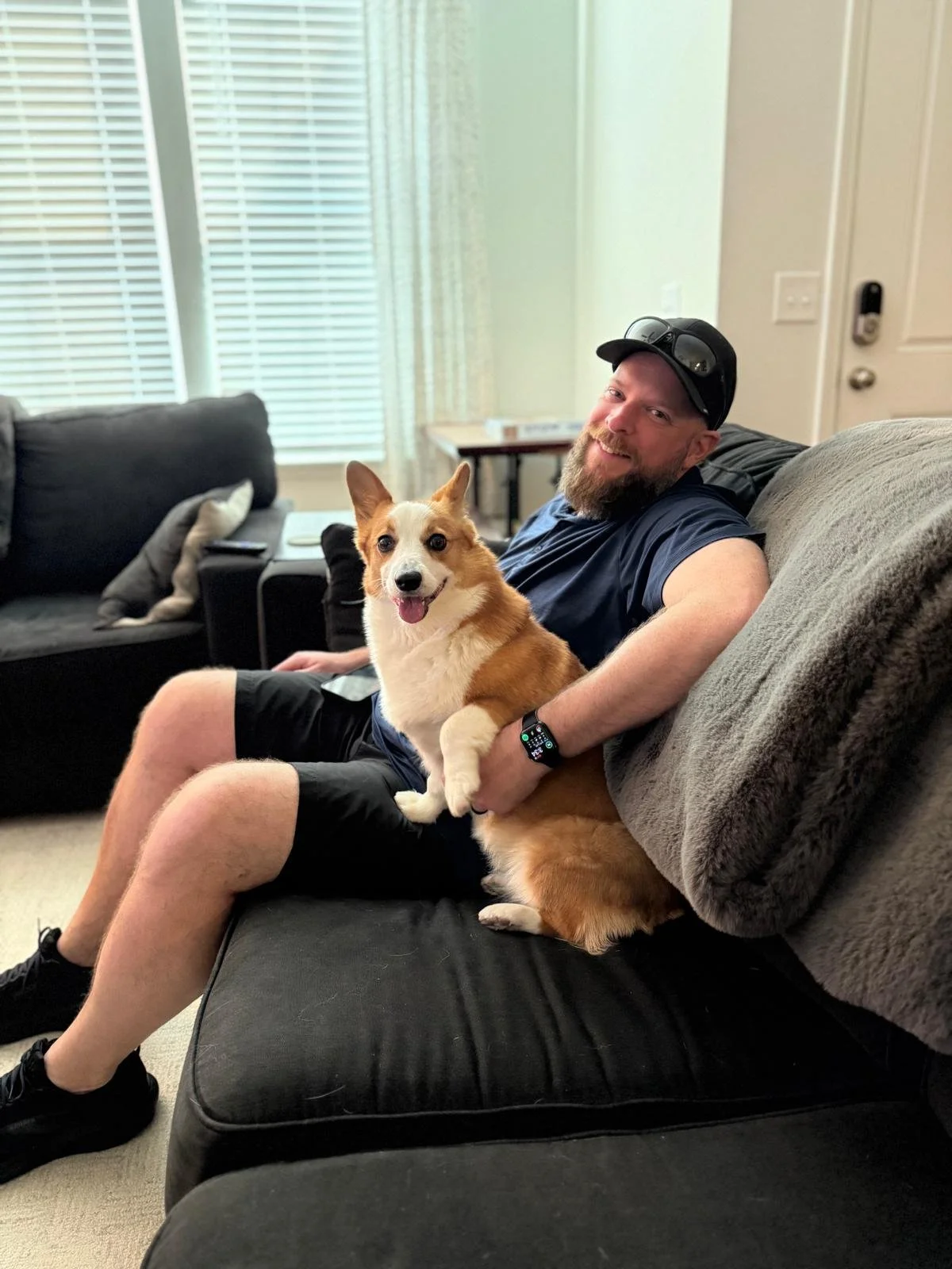 A man with a beard and a cap sitting on a couch, holding a happy Corgi dog, in a living room with large windows with blinds and a table in the background.