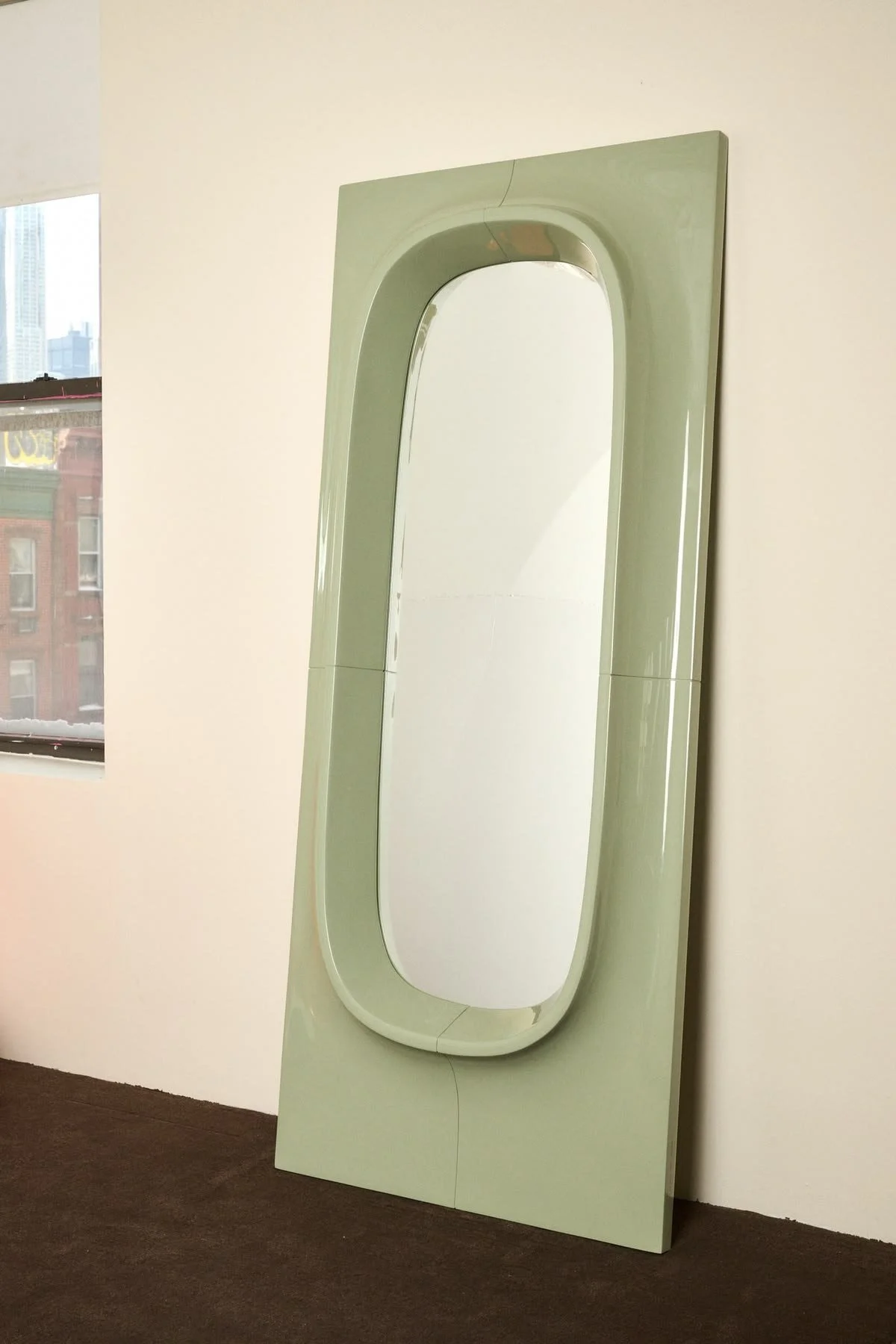 A large, decorative mirror with a light green frame leaning against a white wall near a window in a room.