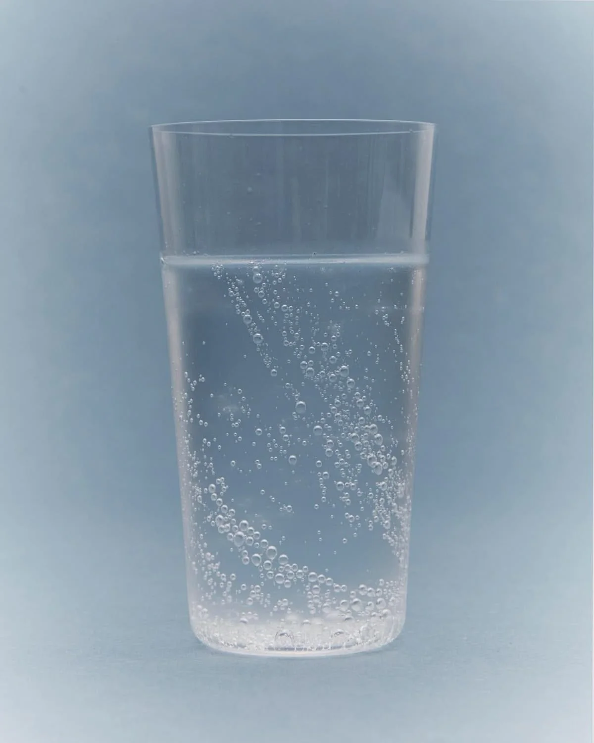 A clear glass filled with carbonated water with bubbles rising inside, set against a light blue background.