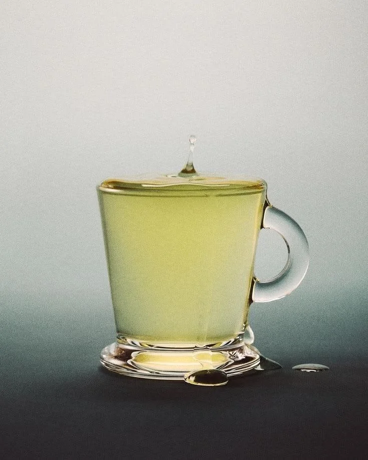 Close-up of a transparent glass cup filled with a light yellow liquid, with a drop of liquid falling into it and spilling over the rim, on a dark surface.