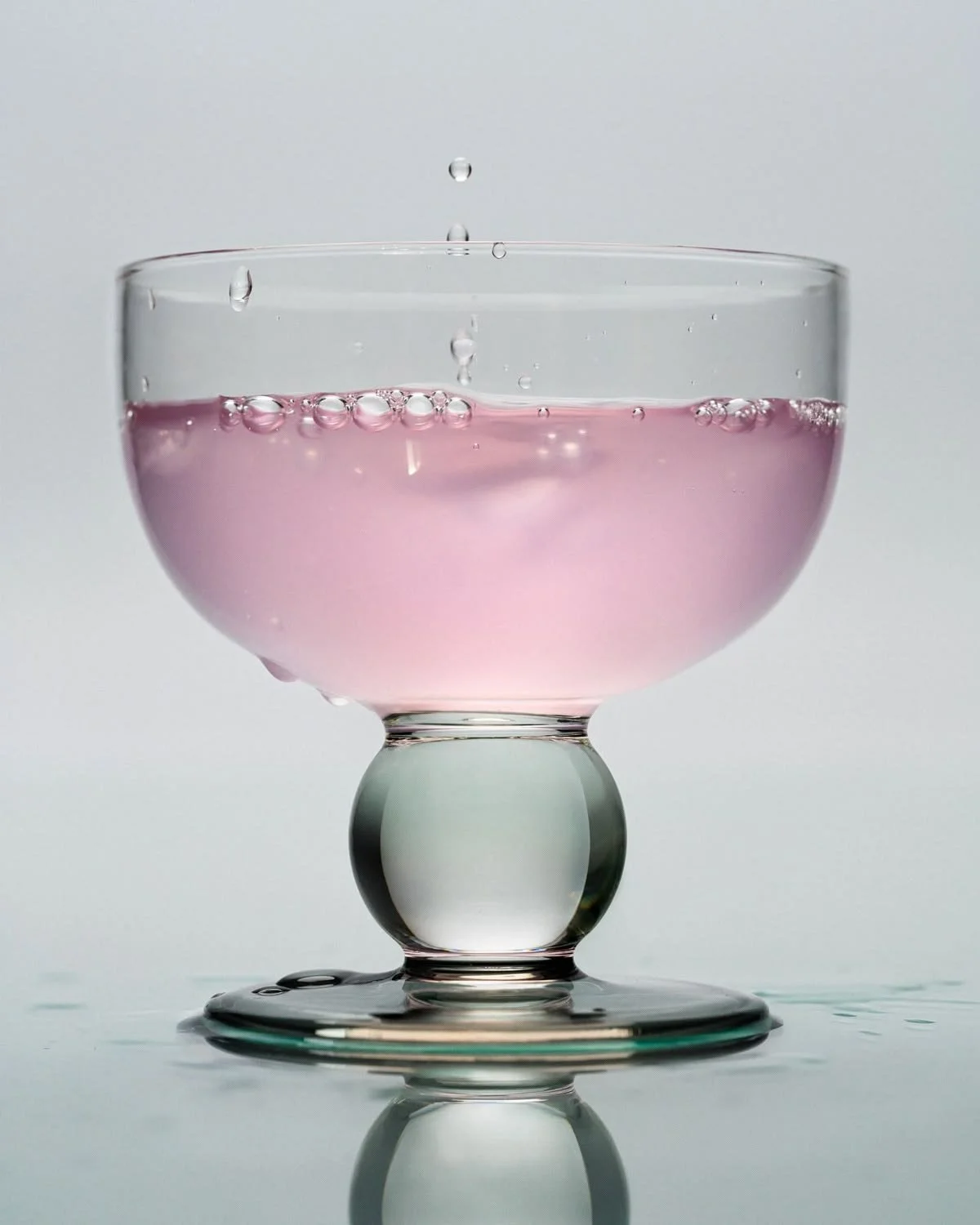 Close-up of a glass filled with pink liquid, with bubbles on the surface and a water droplet falling into it.
