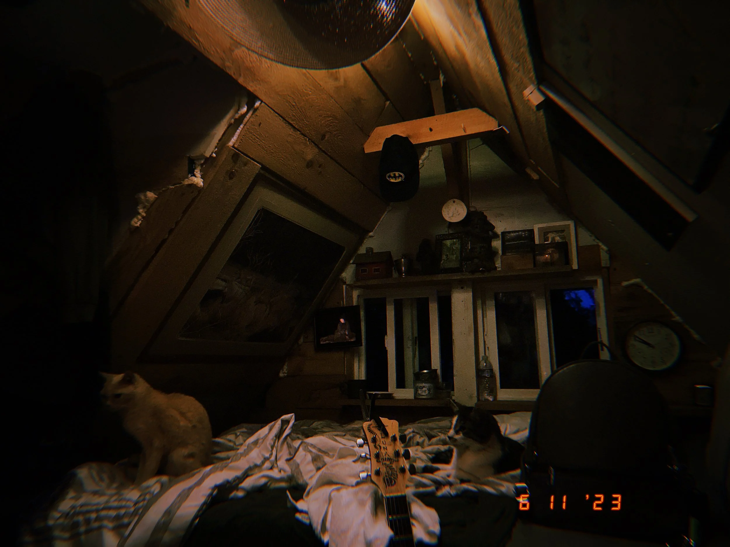Living room at night with a guitar on a crumpled sheet, a cat on the left, and home decor including a baseball cap, dolls, and framed photos. The photo has a timestamp of November 23, 2023.
