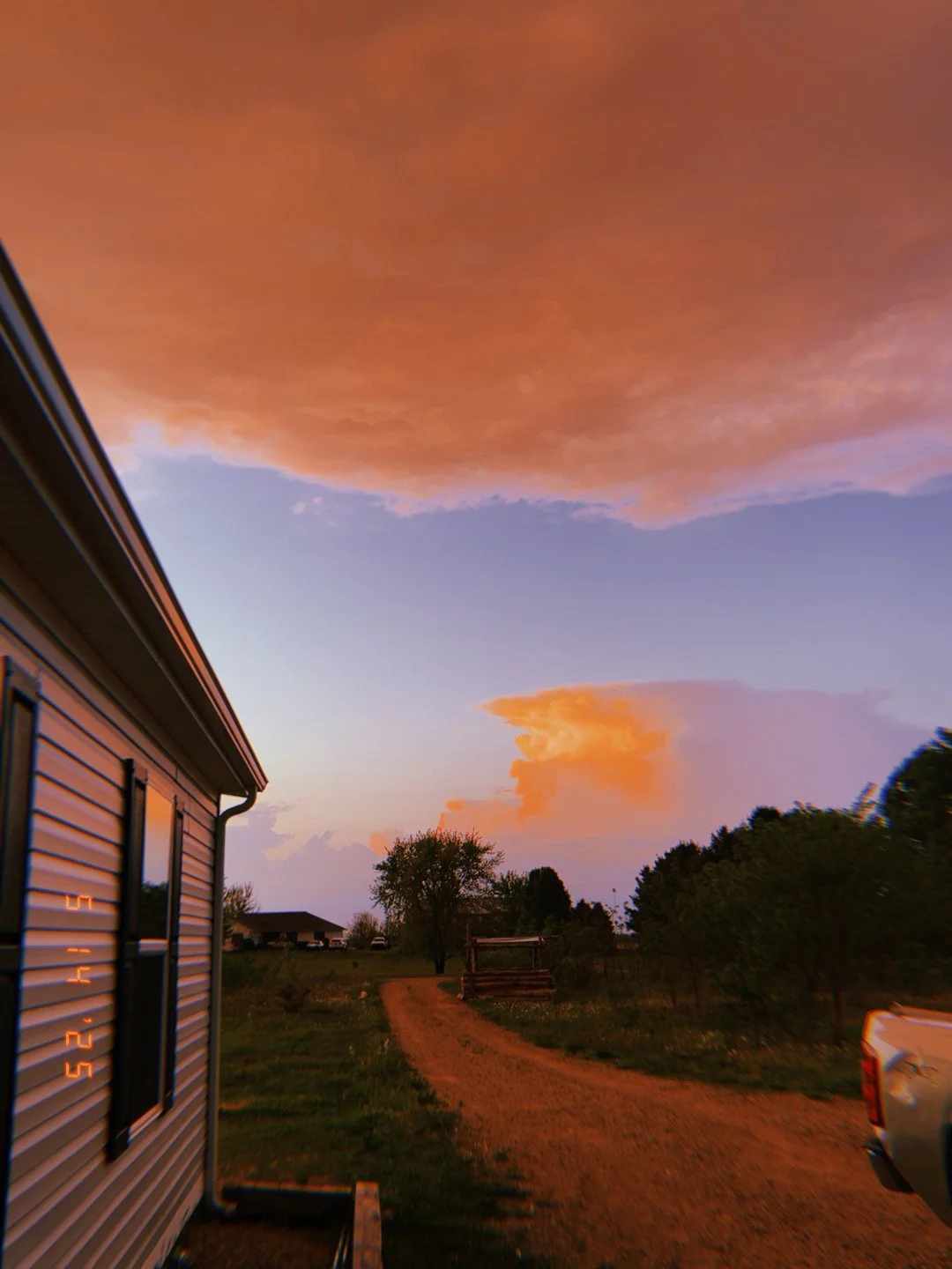 A rural scene during sunset with large orange clouds in the sky, a dirt road, a house on the left, and trees on the right.