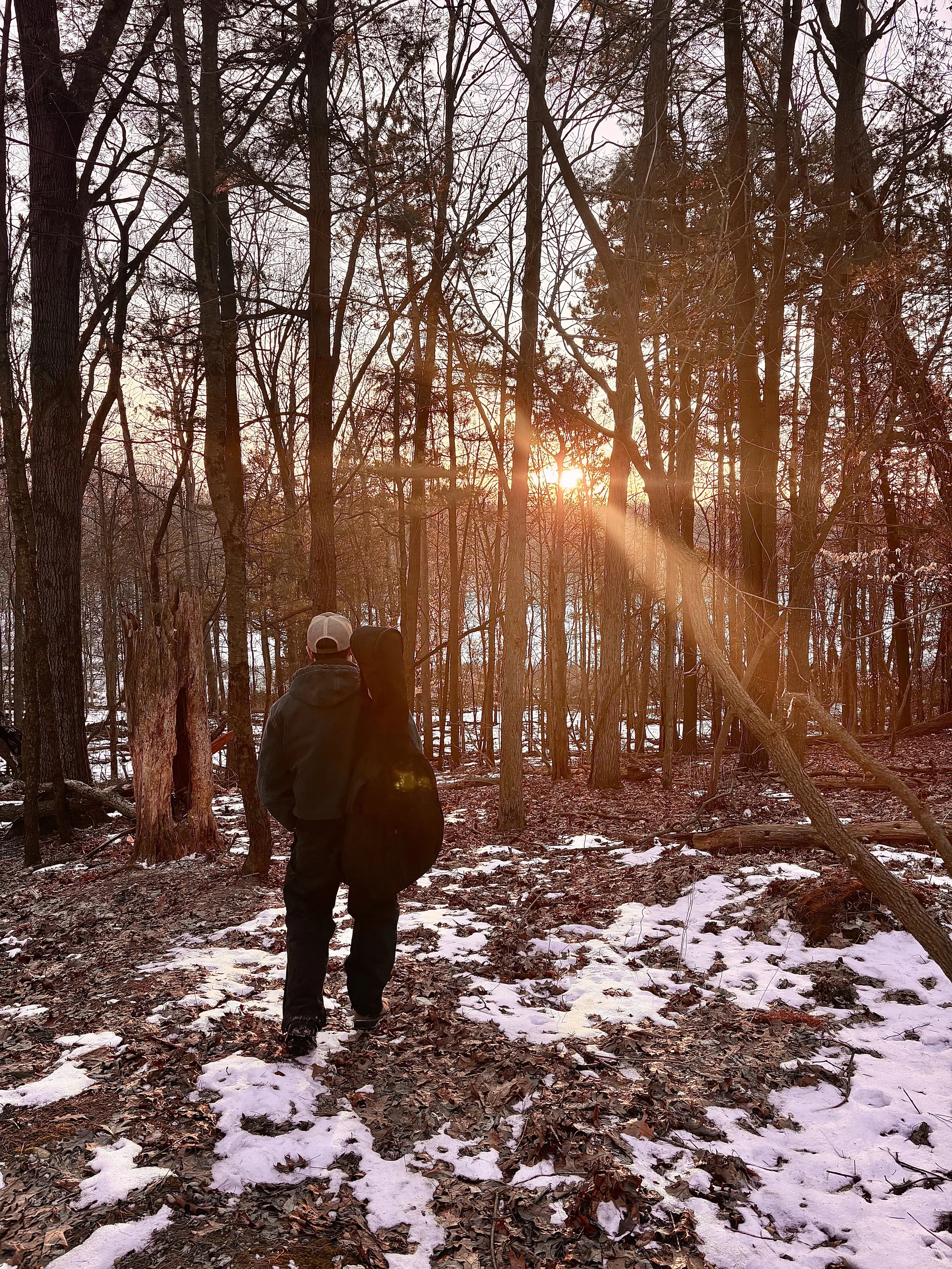 A person walking in a snow-dusted forest during sunset. The person is wearing a gray hoodie and carrying a black bag over their shoulder, and is walking on a path covered with fallen leaves and patches of snow, surrounded by tall trees with bare branches.