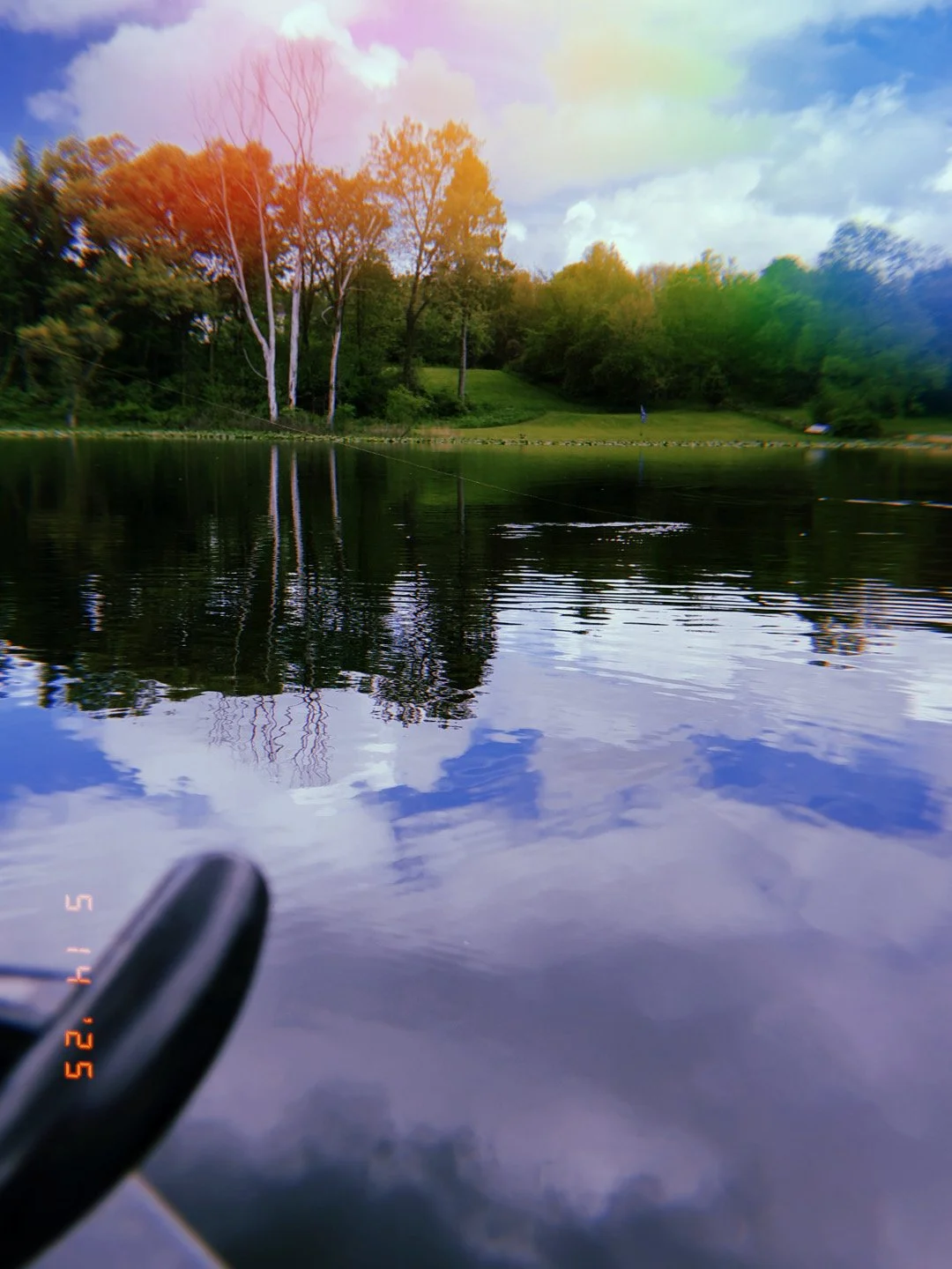 A peaceful lake scene with calm dark water reflecting the trees and sky. In the background, lush green trees and a grassy hillside are visible under a partly cloudy sky. Some trees have bare branches, indicating early spring or fall. The edge of a bo
