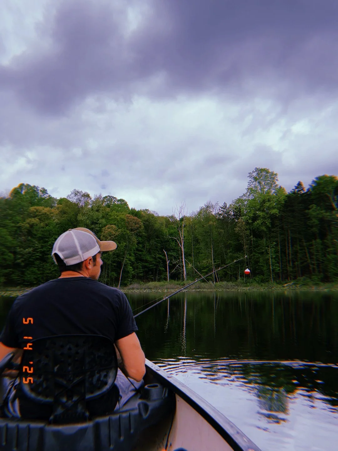 Person fishing from a boat on a calm lake with a forested shoreline and cloudy sky.