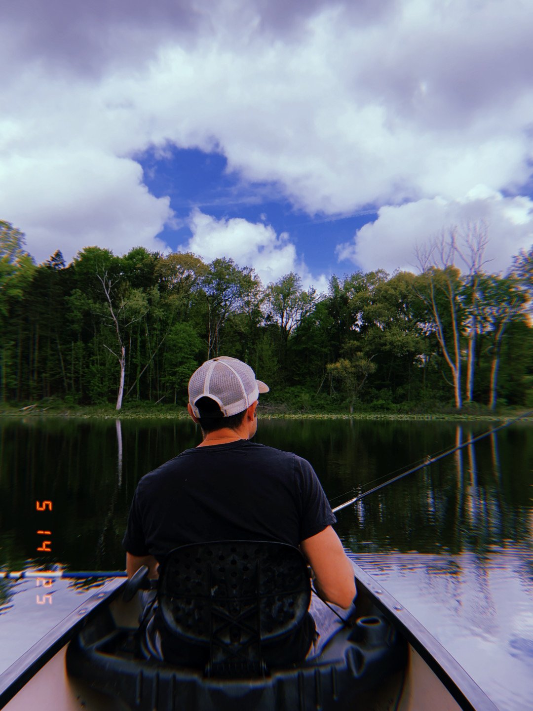 A person wearing a gray trucker hat and black shirt sitting in a boat on a calm lake, fishing with a rod. The background features a treeline with green and some leafless trees under a partly cloudy sky.