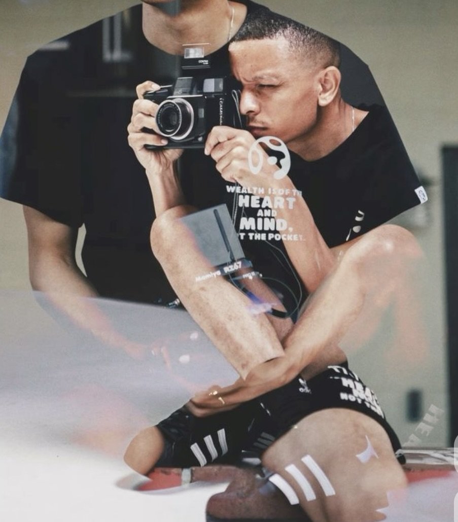 A man taking a photo with a camera in front of a mirror, wearing athletic shorts, with reflections of motivational text on the mirror, including 'Wealth is of the heart and mind, not the pocket' and other words about focus and wealth.