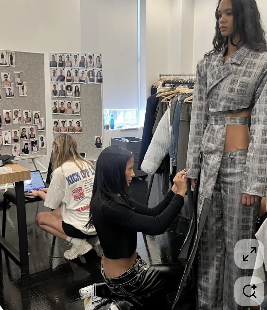 Fashion designer fitting a plaid suit on a mannequin in a studio, with two women working at a computer and looking at photos of models on boards in the background.