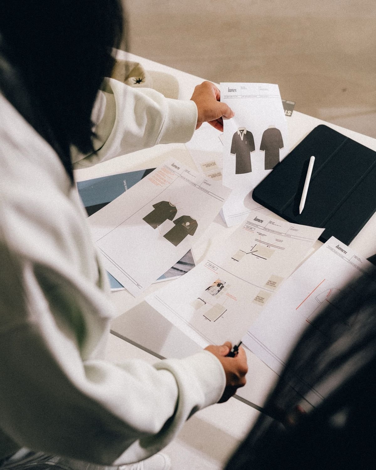 Person looking at clothing design sketches and technical sheets on a desk, with a black notebook and stylus present.
