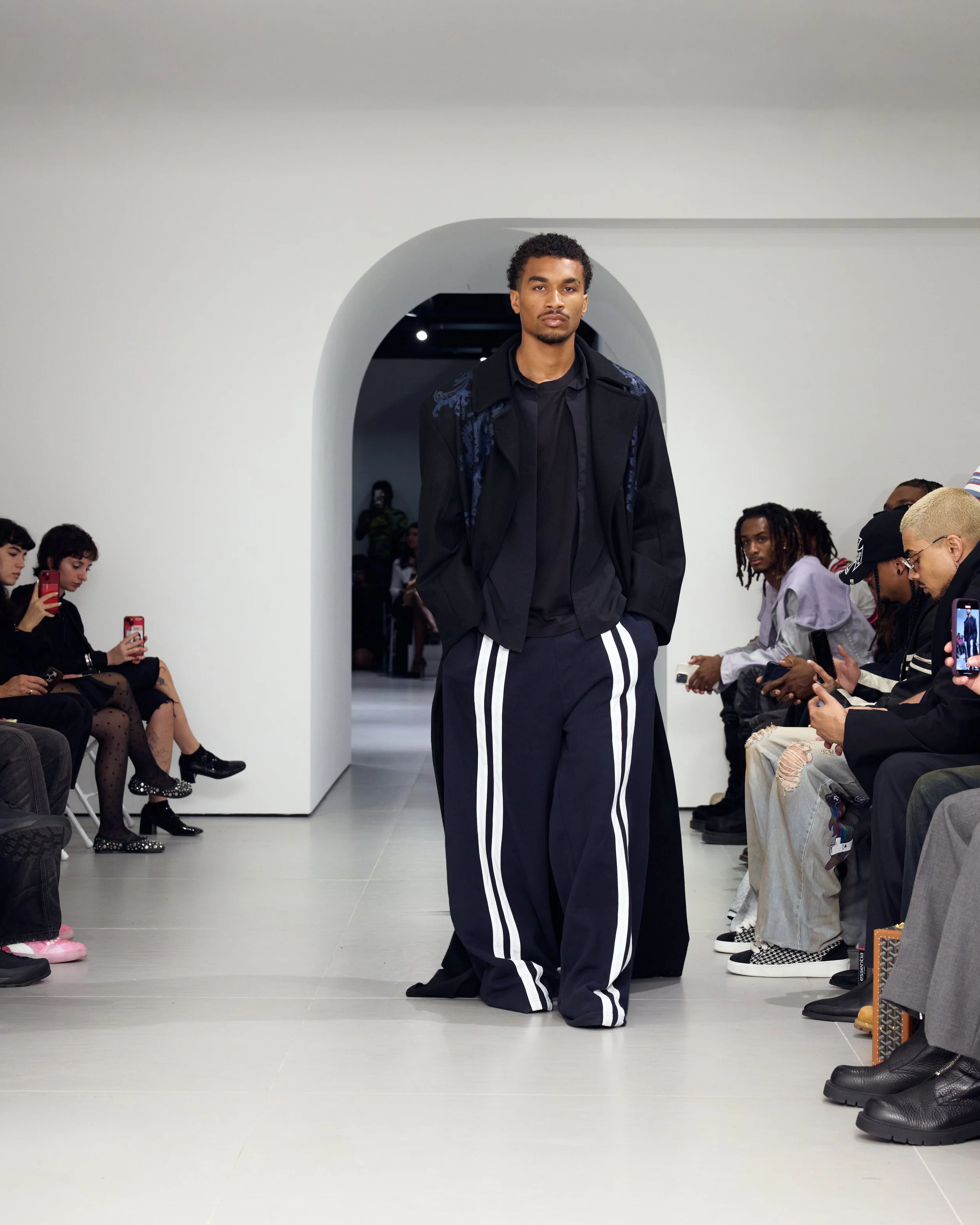 A male model walking down the runway at a fashion show, wearing a black jacket and black pants with white stripes. Audience members are seated on either side, some taking photos with phones or tablets.