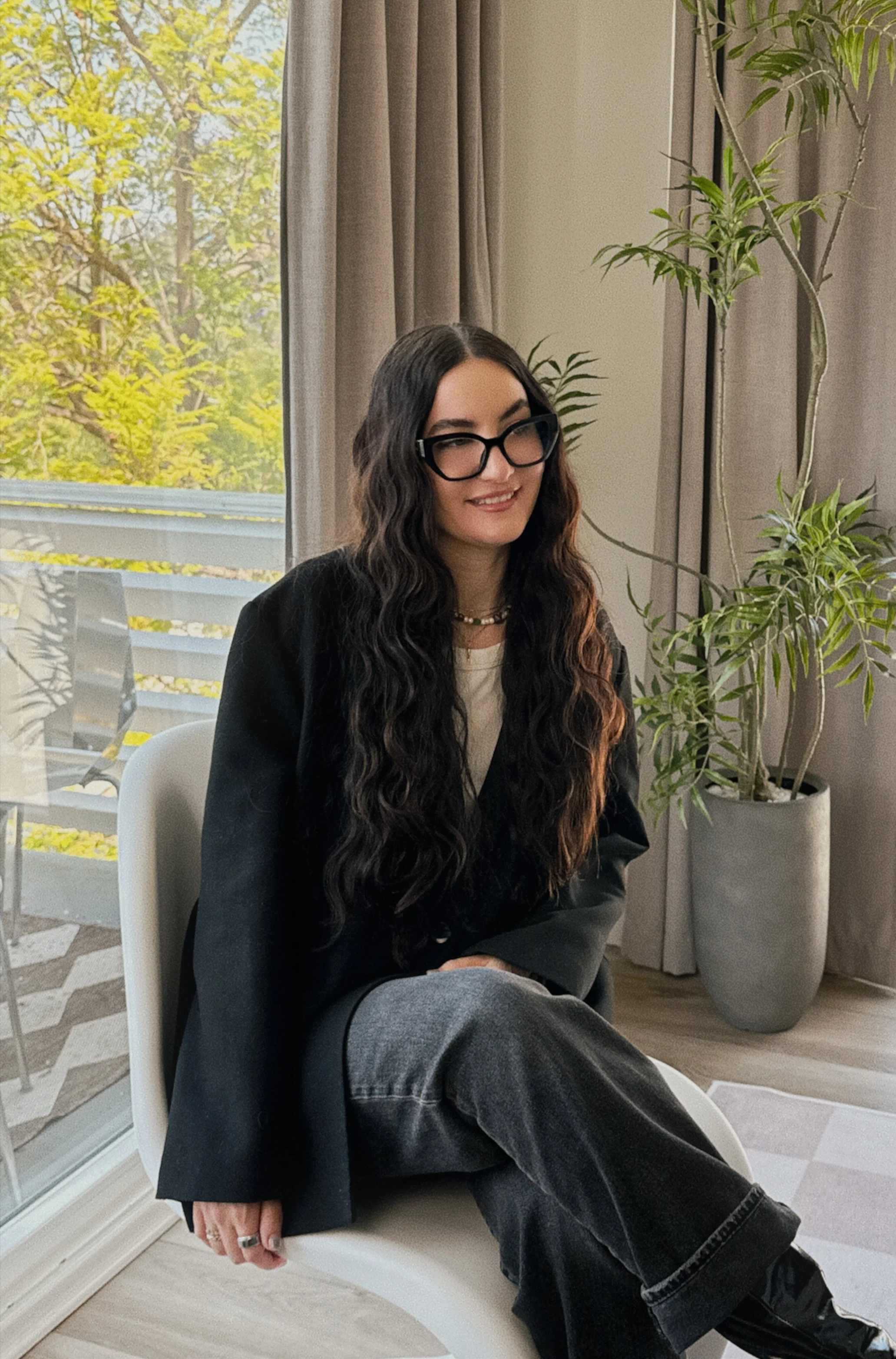 A woman with long wavy dark hair, black glasses, a black blazer, white shirt, and grey jeans, sitting on a white chair indoors. She is smiling and wearing a necklace and ring, with a large potted plant beside her and a window with beige curtains behind her showing a tree outside.