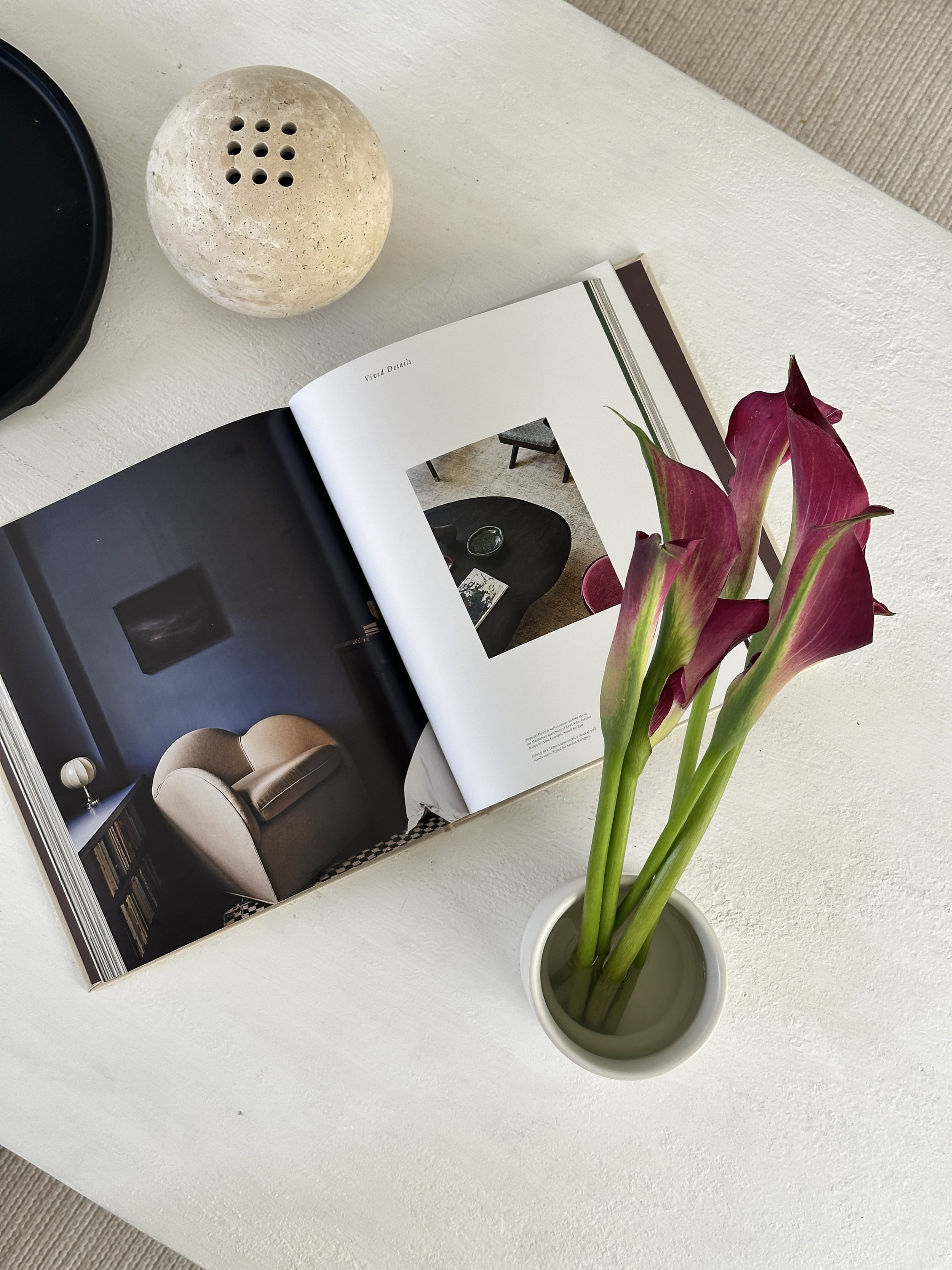 A white table with a magazine open to a page showing a living room, a white vase with pink calla lilies, and a round stone diffuser on the table.