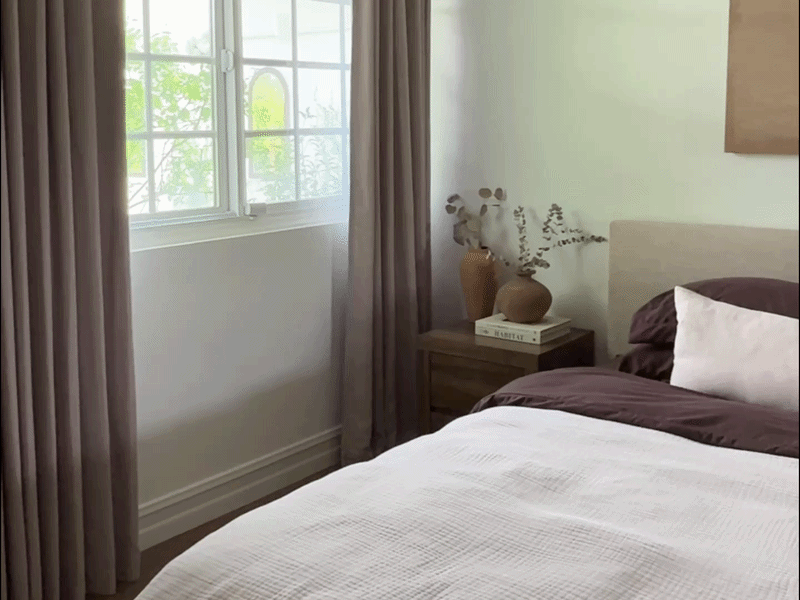 A cozy bedroom with a window, beige curtains, a couch, and a wooden side table with plants and books.