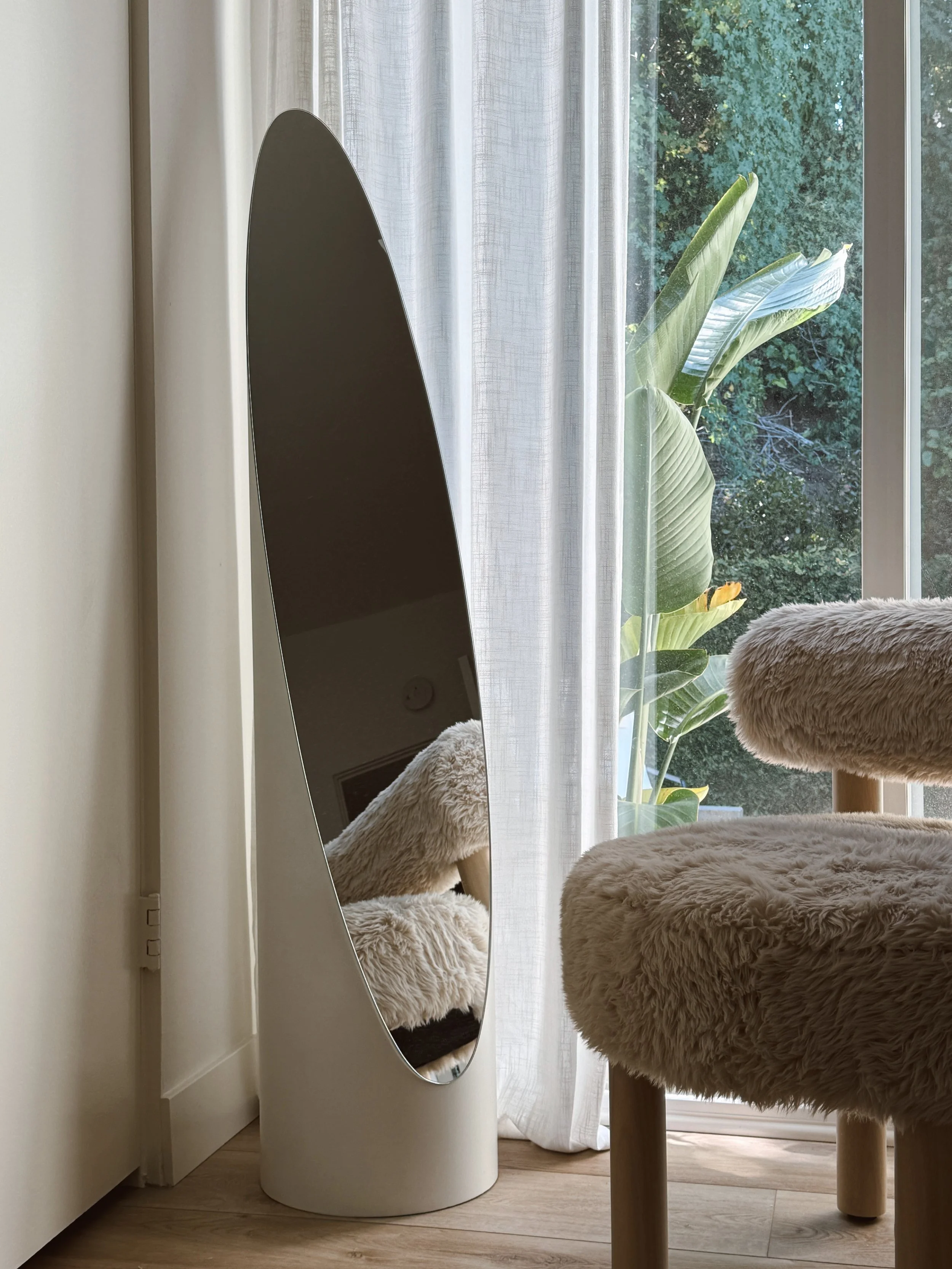 A cozy indoor scene with a fluffy beige armchair and a window with white curtains, revealing green plants outside.