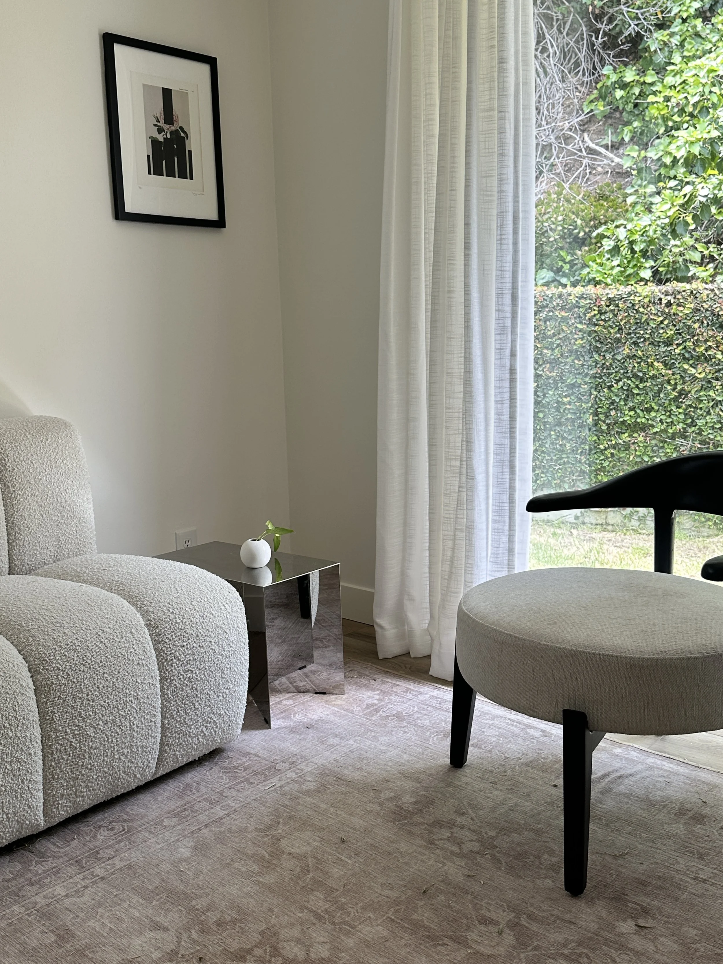 A cozy living room featuring a cream-colored textured armchair, a small reflective side table with a white vase and green plant, a large window with sheer white curtains revealing greenery outside, and a framed botanical print on the wall.