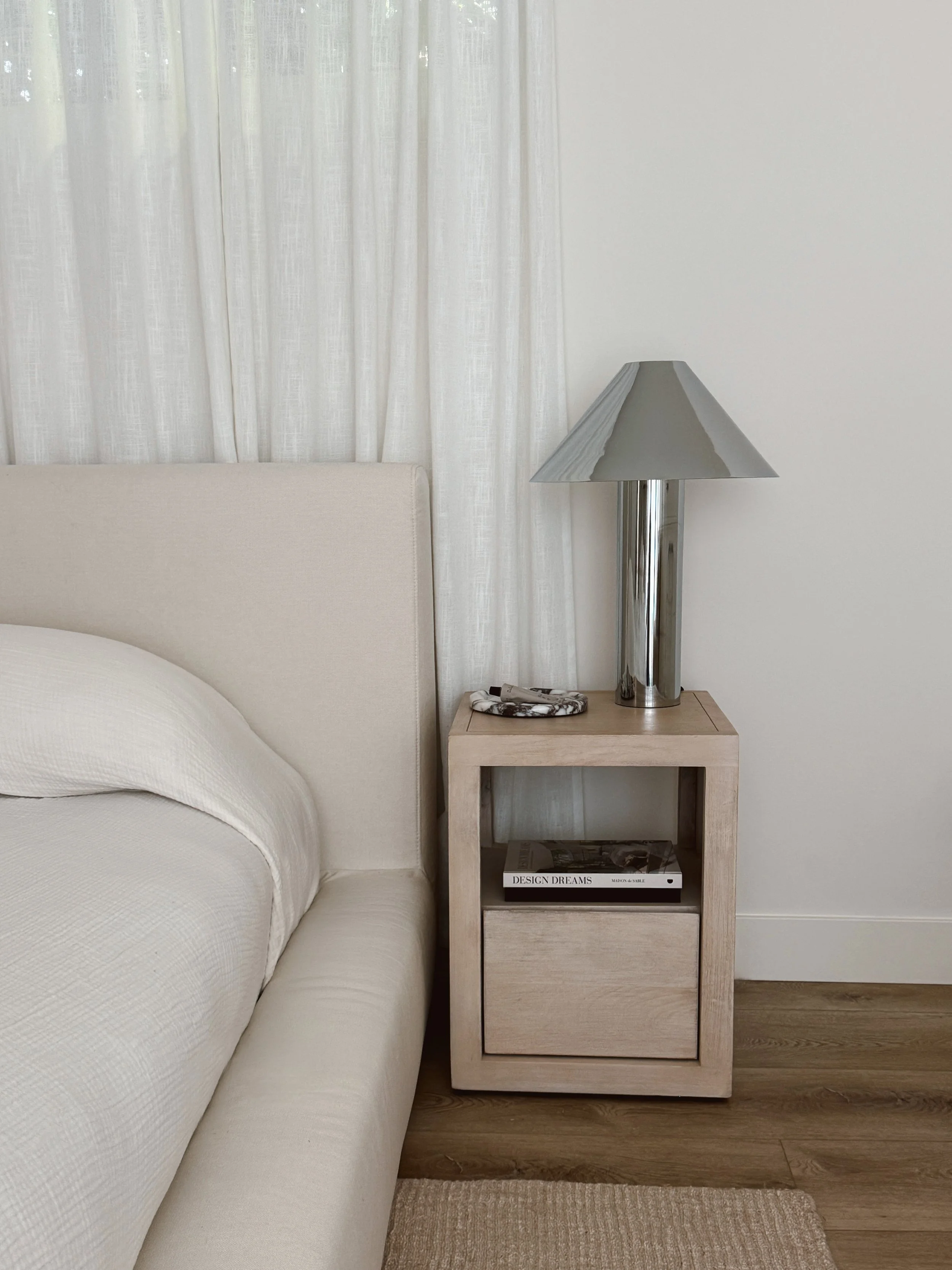 A beige bed with a rounded corner and white textured bedding, next to a light wood nightstand with a single drawer and an open shelf. The nightstand holds a modern silver lamp with a cone-shaped shade and a decorative tray. Books are on the shelf, titled 'Design Dreams' and 'Maison de Sable.' The background features a white wall with sheer white curtains, and the floor is light wood with a beige rug.