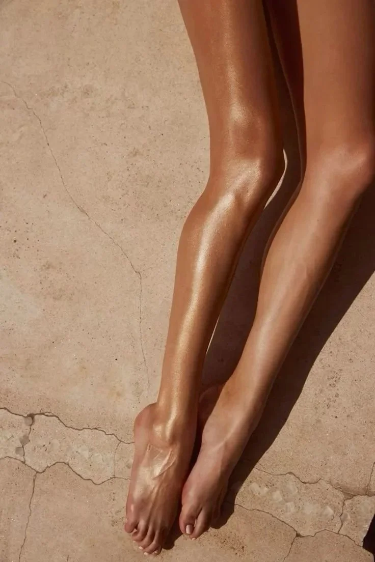 Close-up of two shiny, brown, and tan human legs with smooth skin, lying on a cracked, beige ground.
