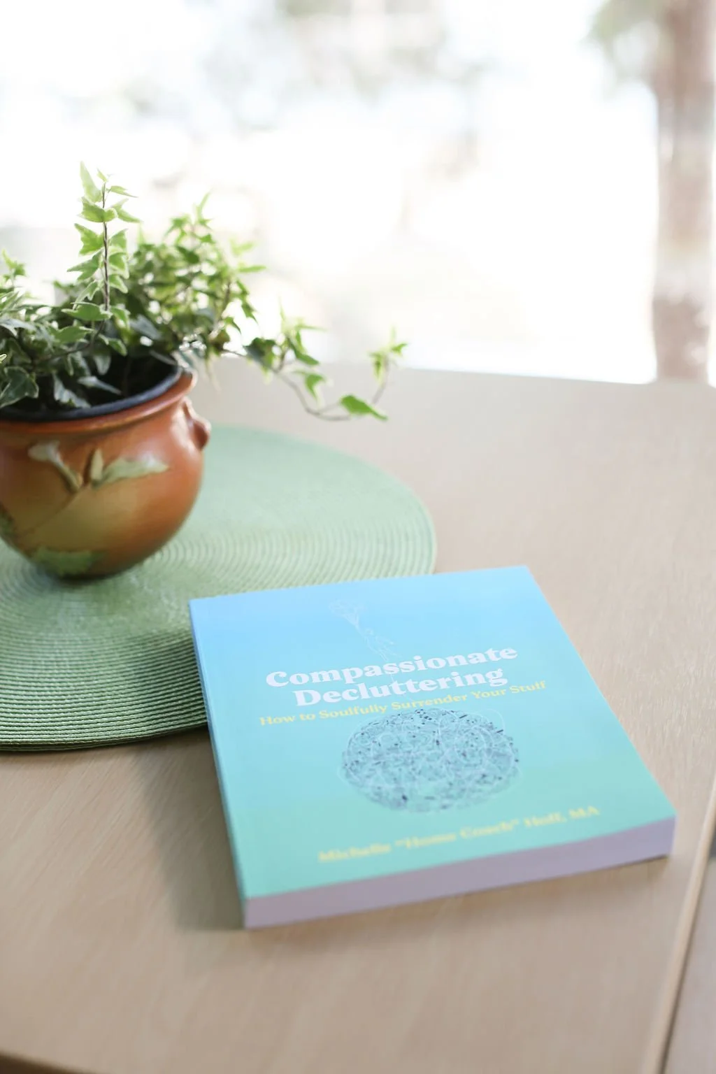 A copy of the Compassionate Decluttering book on a table beside a potted plant.