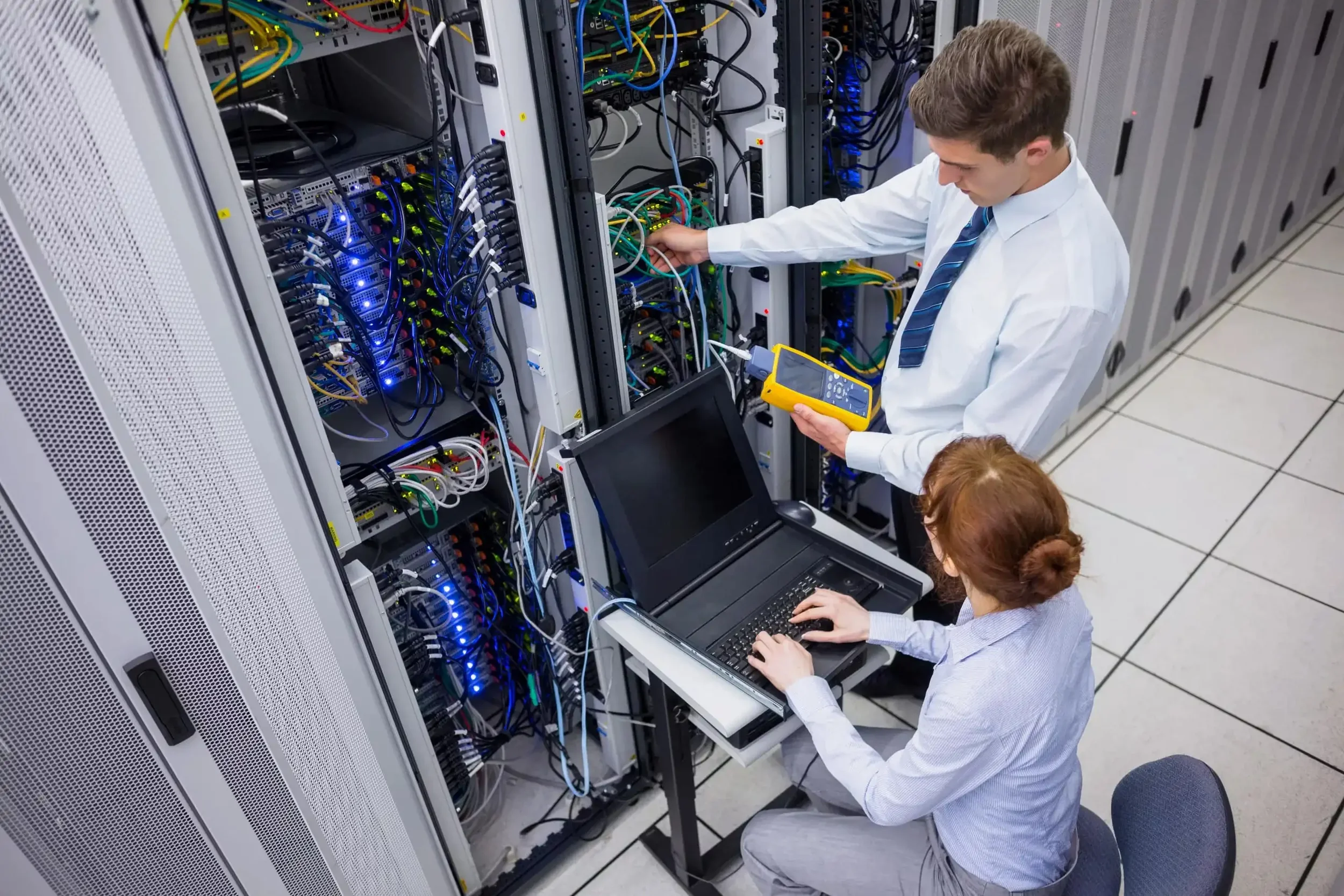 Two women working on a computer server rack in a data center. One woman is seated at a laptop, and the other woman is standing and adjusting cables in the server rack.