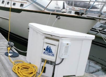 A white boat dock utility box with a yellow garden hose coiled beside it, located at a marina with other boats in the background.