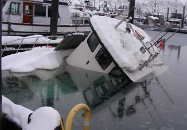 A boat partially submerged and tilted in icy water at a marina during winter, covered with snow.   MarCELL PRO would prevent this from happening by alerting the owner that high water in the bilge has been detected