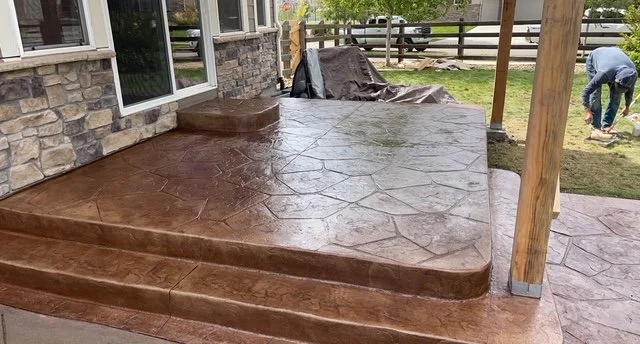 Newly stained concrete patio with curved steps outside of a house, with a man in the background working on the yard.