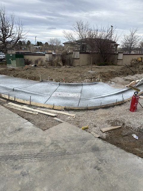 Construction site with curved concrete sidewalk or pathway being built, surrounded by construction materials and equipment, including a fire extinguisher.