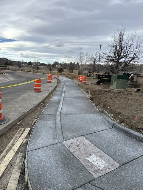 Newly paved sidewalk under construction with orange traffic barrels and caution tape, cloudy sky, trees, and street in the background.