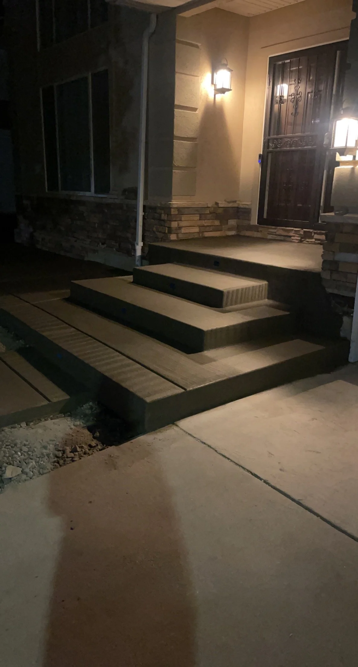 Nighttime photo of a house entrance with steps, a door with a wrought iron security gate, a wall-mounted lantern light, and a porch area with stone and wood elements.