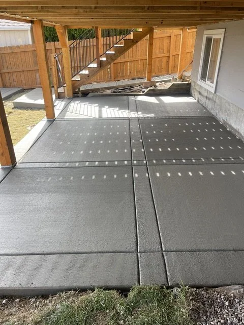 Newly poured concrete patio with a wooden deck above, wooden stairs, and a fenced backyard. Shadows of the deck posts create a dotted pattern on the concrete.