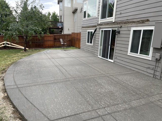 Concrete patio area with a house wall, windows, and a fence in the background, with a basketball hoop on the fence.