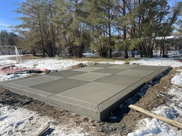 Outdoors scene with a dry, newly poured concrete slab on a snowy ground, surrounded by bare trees and a few distant structures.