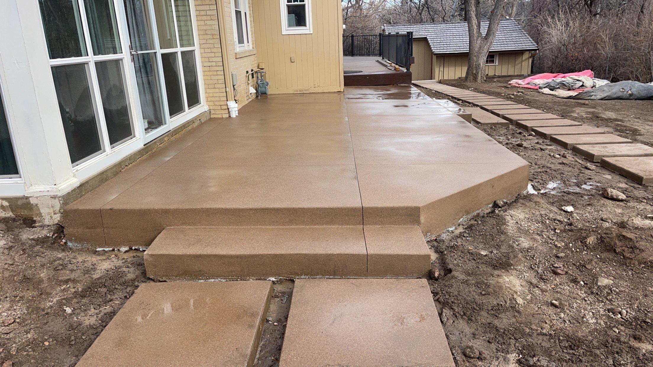 Newly installed concrete patio with steps leading down to bare soil, side of house with large windows, and backyard with dirt, trees, and construction materials.