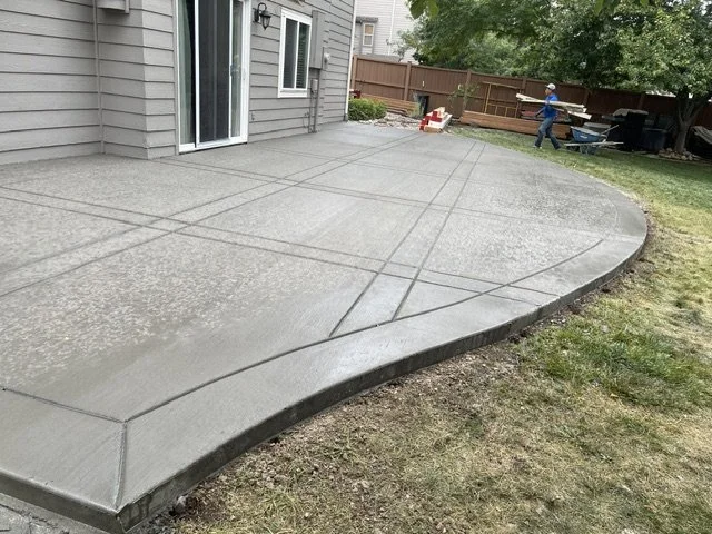 Freshly poured concrete patio with visible expansion joints next to a house, with a person working in the background in a backyard.