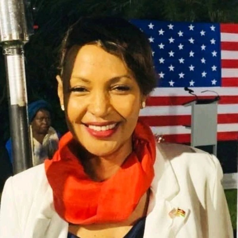 Smiling woman with short dark hair, white blazer, red scarf, and a small American flag pin on her blazer, in front of an American flag backdrop.