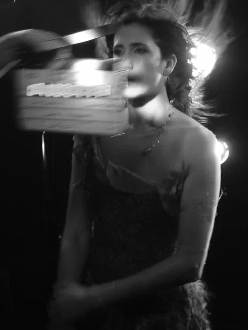 A woman with curly hair and a necklace stands in front of a bright light, while a clapperboard obscures part of her face. The photo is in black and white.